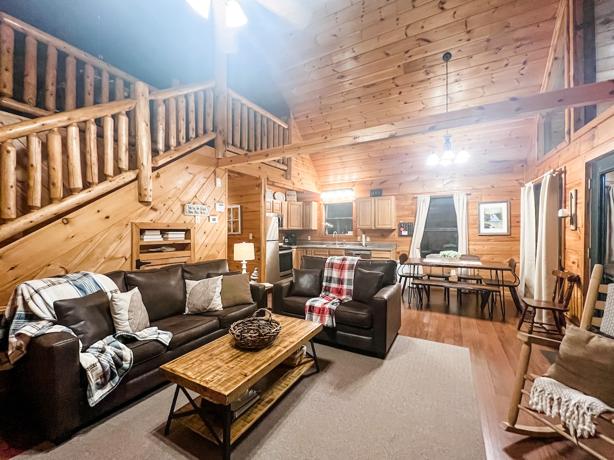 A spacious living room features vaulted wood ceilings and is enhanced by large windows. Comfortable leather sofas, a wooden coffee table, and a cozy throw blanket create an inviting atmosphere. The open layout connects to a dining area and kitchen in the background.