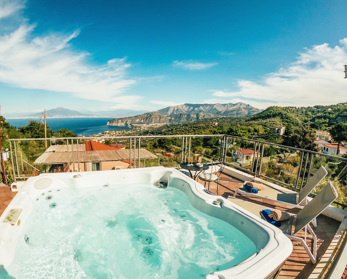 The rooftop terrace features a Jacuzzi surrounded by a glass railing, offering stunning views of the expansive Gulf of Naples and distant mountains. Two lounge chairs sit nearby, inviting relaxation under the clear blue sky.