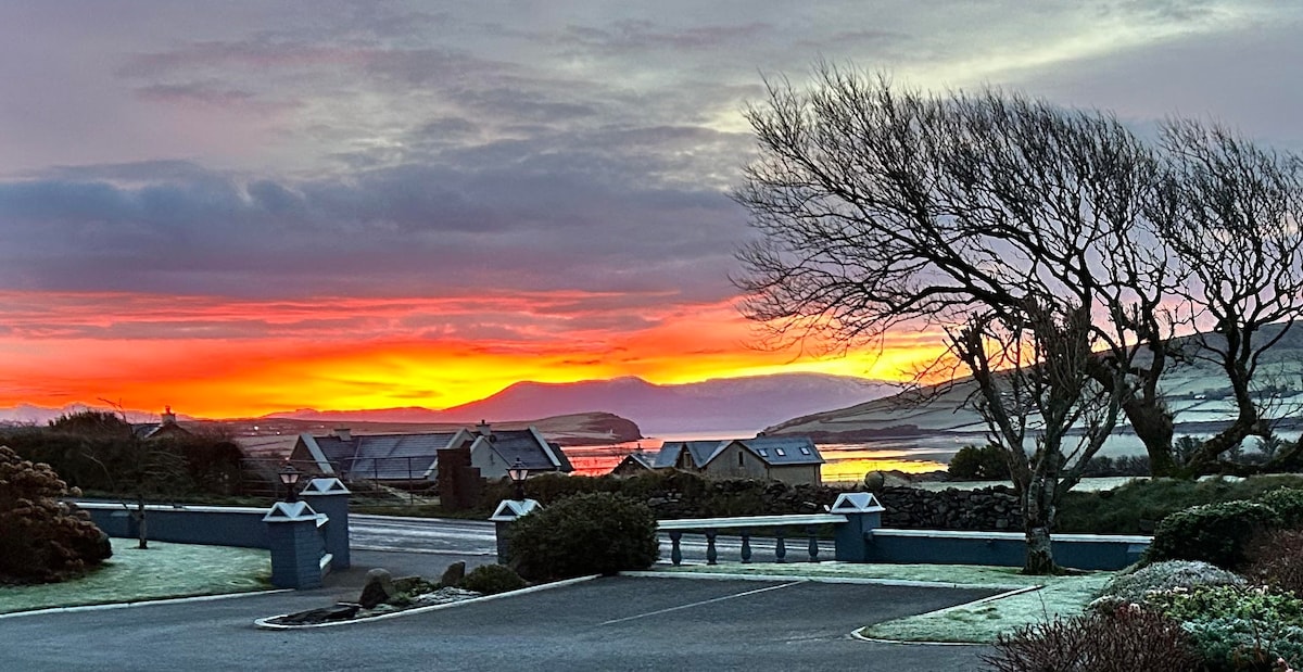 A stunning sunset is observed over a tranquil waterfront, with vibrant hues of orange and pink illuminating the clouds. Silhouetted trees line the foreground, and a collection of cottages is visible in the distance, set against the backdrop of rolling hills.