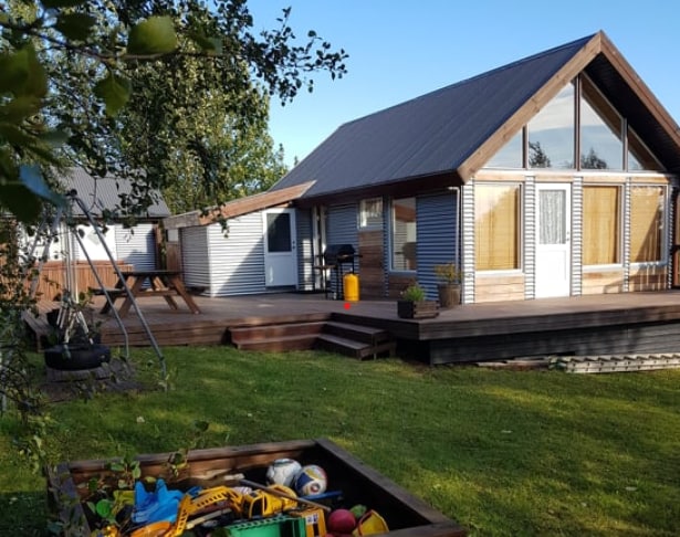 A charming country house is situated in a spacious outdoor area. A wooden deck extends from the house, with steps leading to a lawn filled with greenery. A swing set is visible nearby, and a box containing various outdoor toys is positioned on the ground.