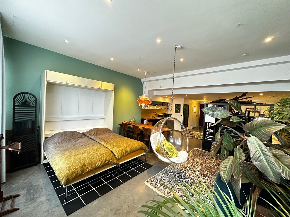 A spacious bedroom showcases a large bed adorned with warm, neutral-toned bedding. A bubble chair is suspended nearby, providing a unique seating option. The walls feature a calming green hue, with a dining area observed in the background, seamlessly integrating the living space.