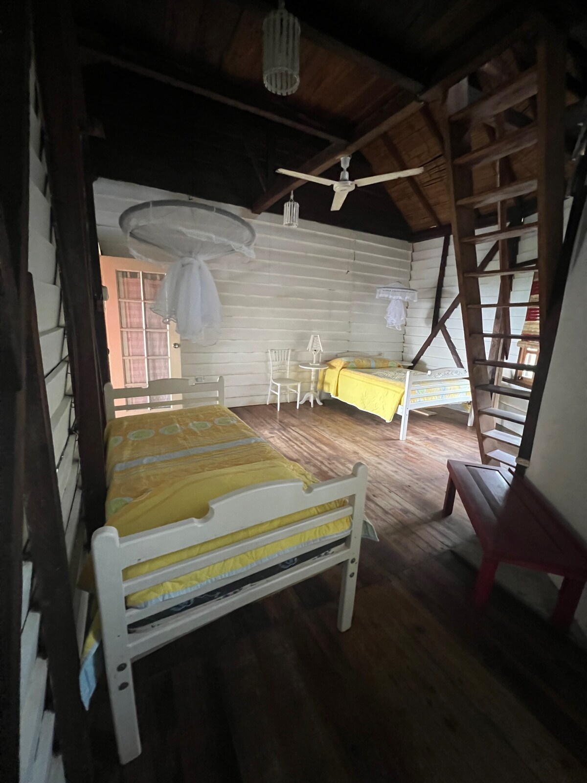 A cozy private room is seen, featuring light wooden walls and a warm wooden floor. The room includes a Queen bed and two twin beds, all dressed in yellow bedding. A ceiling fan is visible overhead, and a staircase leads to an upper area.