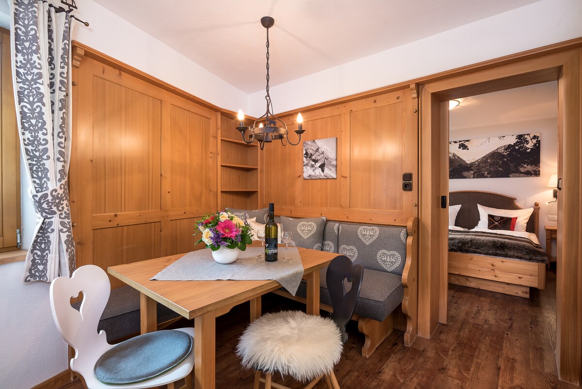 A wooden dining area features a sturdy table surrounded by cushioned benches and a decorative chair. A vase with colorful flowers sits on the table, while a cozy blanket lays over one of the benches. Soft lighting is provided by a chandelier, and a doorway leads to a sleeping area.