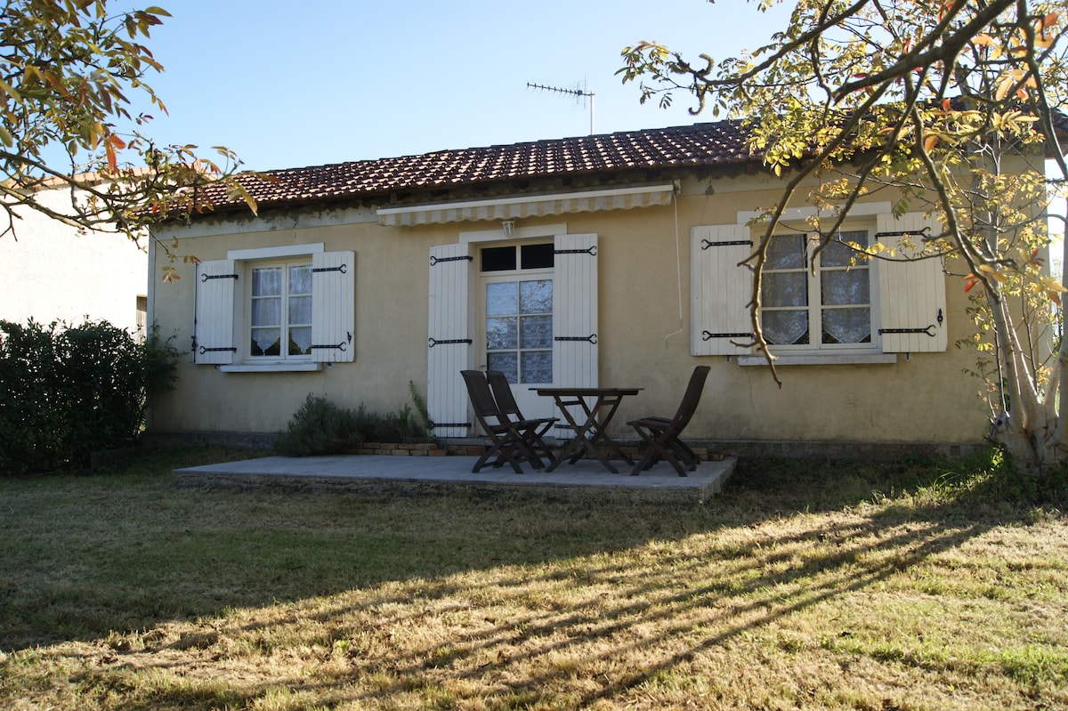 A quaint house is set in a grassy area, featuring a patio with a wooden table and two chairs. The exterior showcases light-colored walls with white shutters and window frames. Sunlight casts shadows from nearby trees, creating a calm outdoor space.