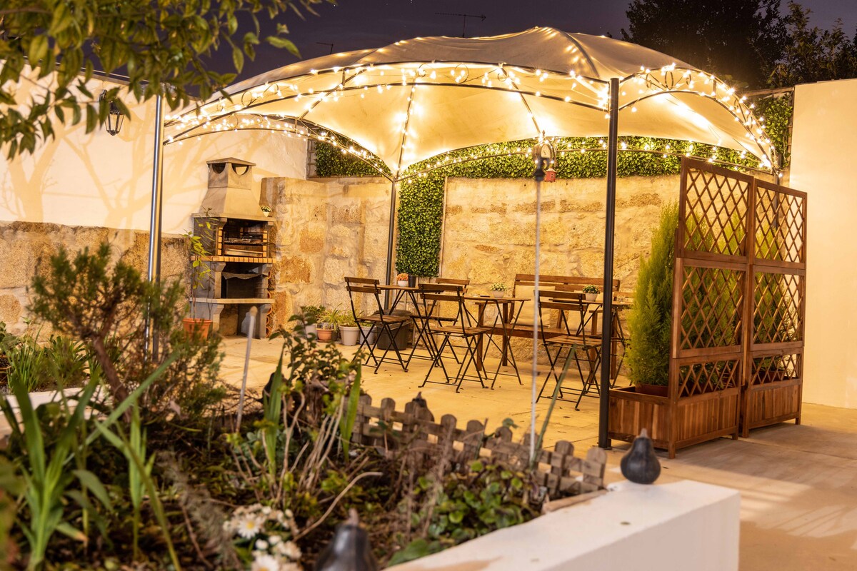 An outdoor dining area features a canopy adorned with string lights, casting a warm glow. Simple wooden tables and chairs are arranged beneath it, while a stone barbecue sits against a textured wall. Lush greenery surrounds the space, adding to the natural ambiance.