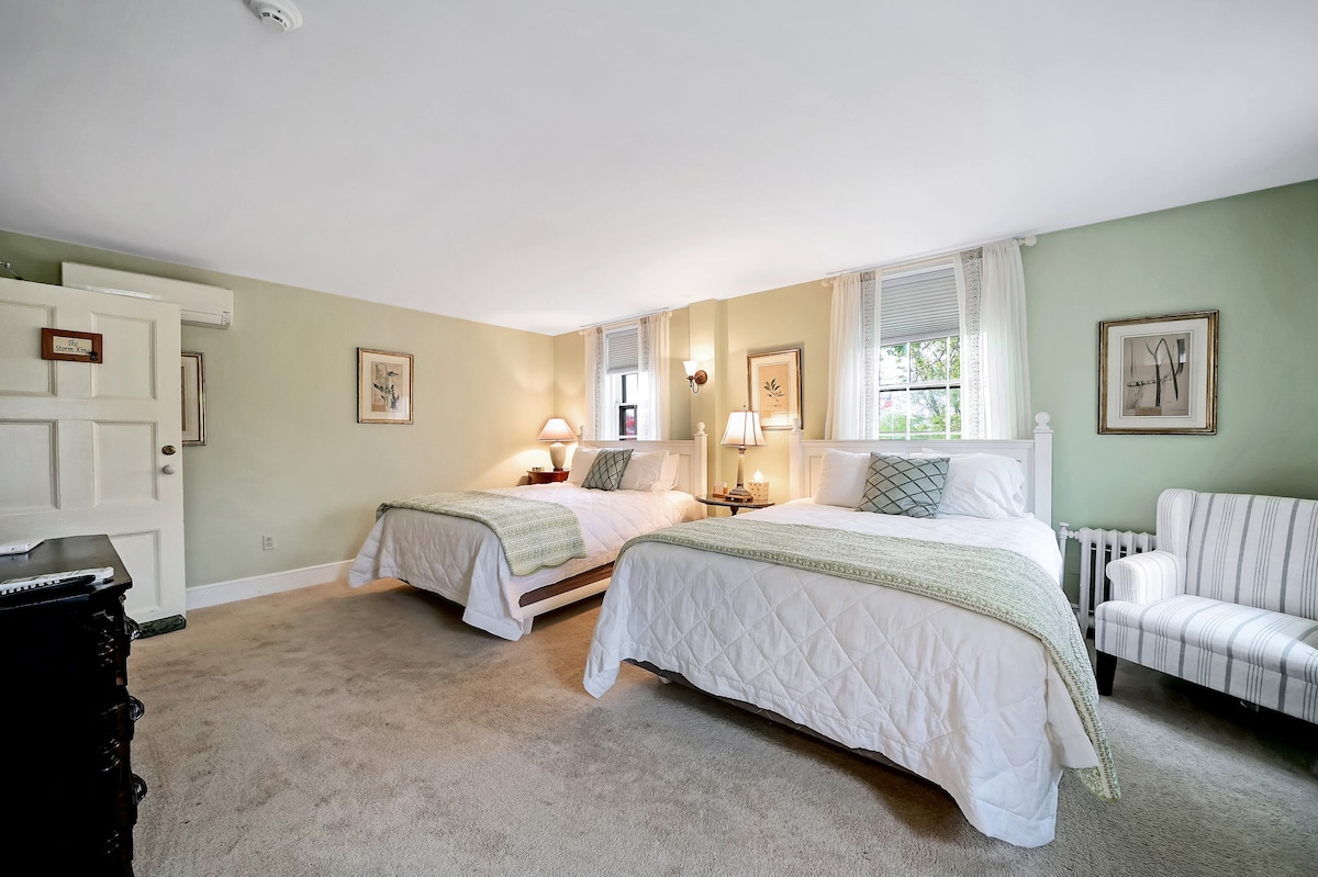 The room features two spacious beds with light-colored bedding against sage green walls. Natural light filters through multiple windows, illuminating the cozy armchair and antique dresser. A door leads to the hall, enhancing the room's accessibility.