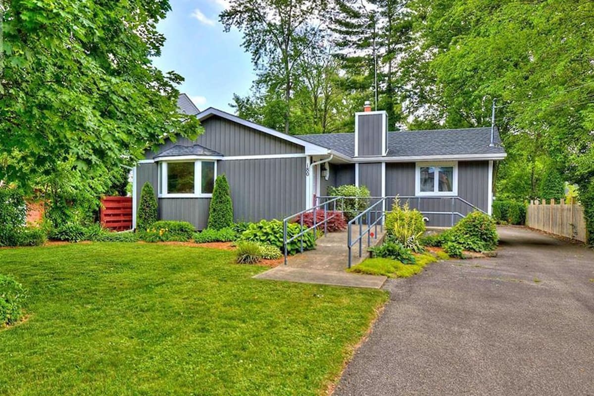 A single-story home is set amidst lush greenery, featuring a manicured lawn and a paved pathway leading to the entrance. A ramp provides accessible access, while various trees and shrubs enhance the natural surroundings.