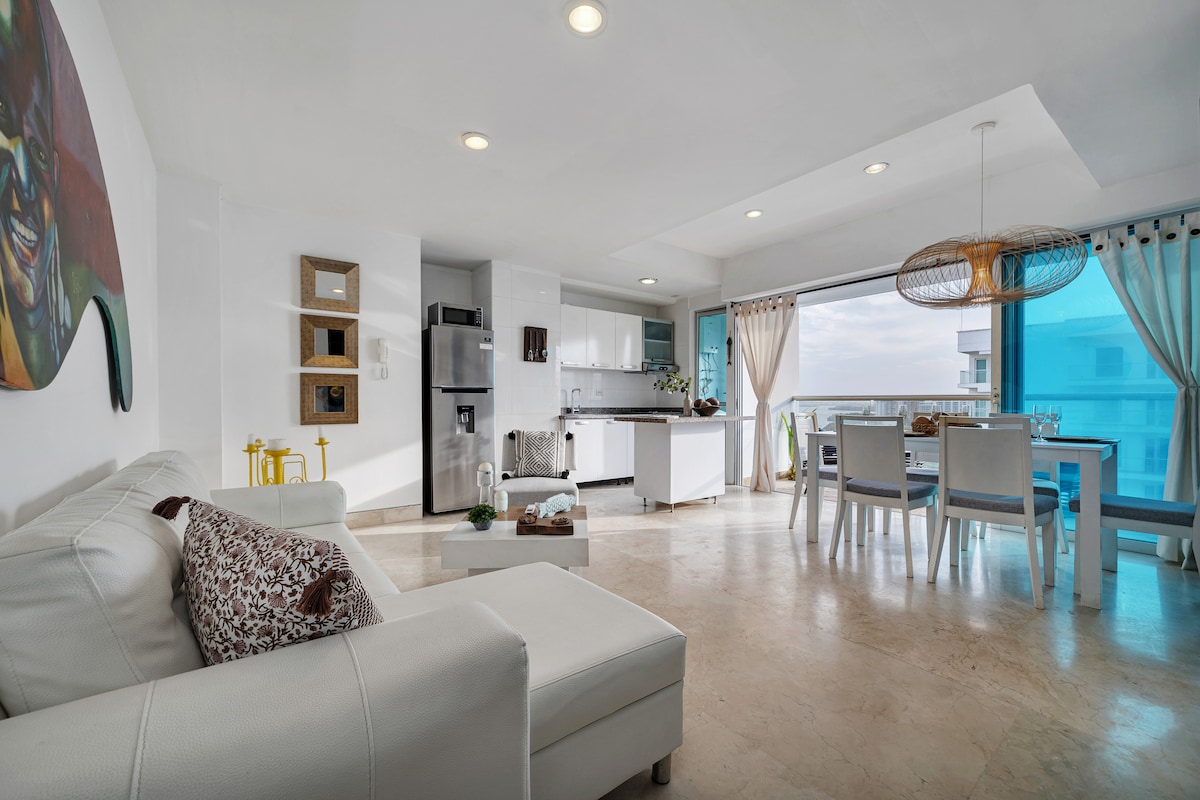 A bright and airy living space features a white sectional sofa and a dining area with a table set for four. Modern kitchen appliances are visible, along with large windows showcasing expansive views. The decor includes subtle Caribbean accents, complementing the open layout.