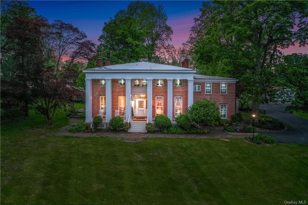 The exterior of a grand two-story brick building is framed by lush greenery. Large columns support the elevated entrance, where warm light spills from the front doors and windows. The manicured lawn extends in front, enhanced by the colors of a sunset sky.