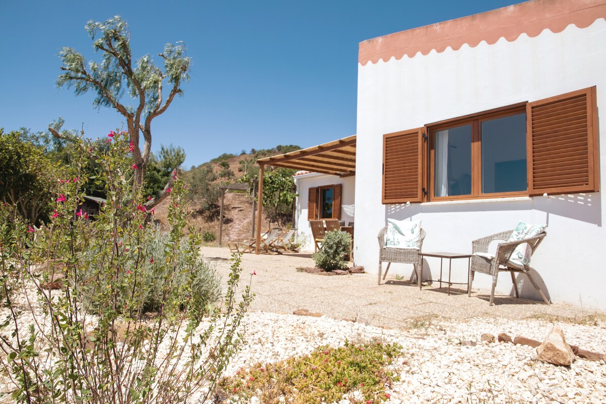 The exterior of the property features a light-colored facade with wooden shutters. Two comfortable chairs are positioned on a patio underneath a pergola. Surrounding greenery and flowering plants create a welcoming atmosphere, with a clear blue sky above.