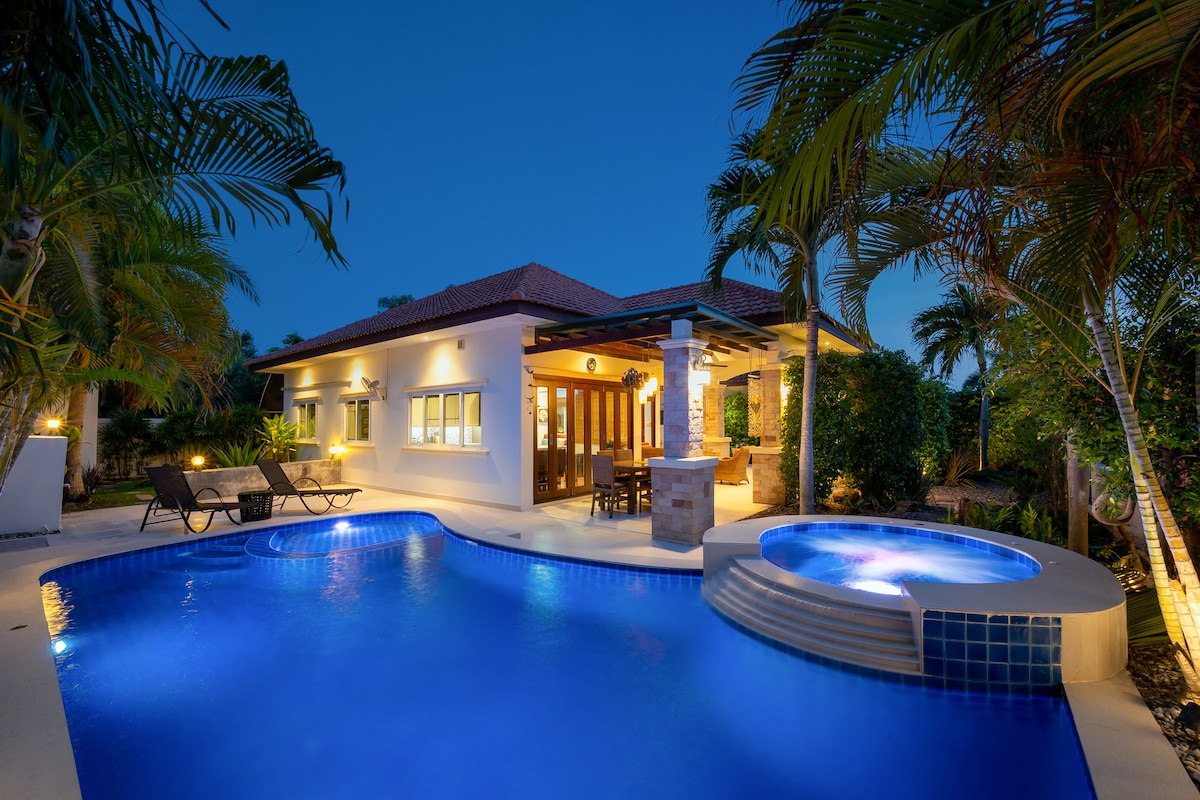 The villa's outdoor area features a saltwater pool with a jacuzzi, surrounded by palm trees. Loungers are positioned beside the pool. The exterior is illuminated softly, enhancing the tranquil atmosphere as evening approaches. The structure includes large windows and a covered seating area.