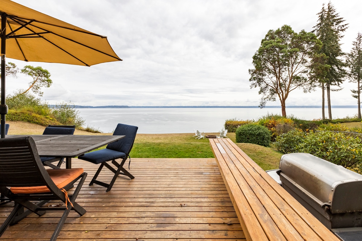 An outdoor deck area features a wooden table set with four chairs beneath a yellow umbrella. A long bench runs alongside the deck, and a grill is positioned nearby. The view extends across calm water and greenery, offering a peaceful natural setting.