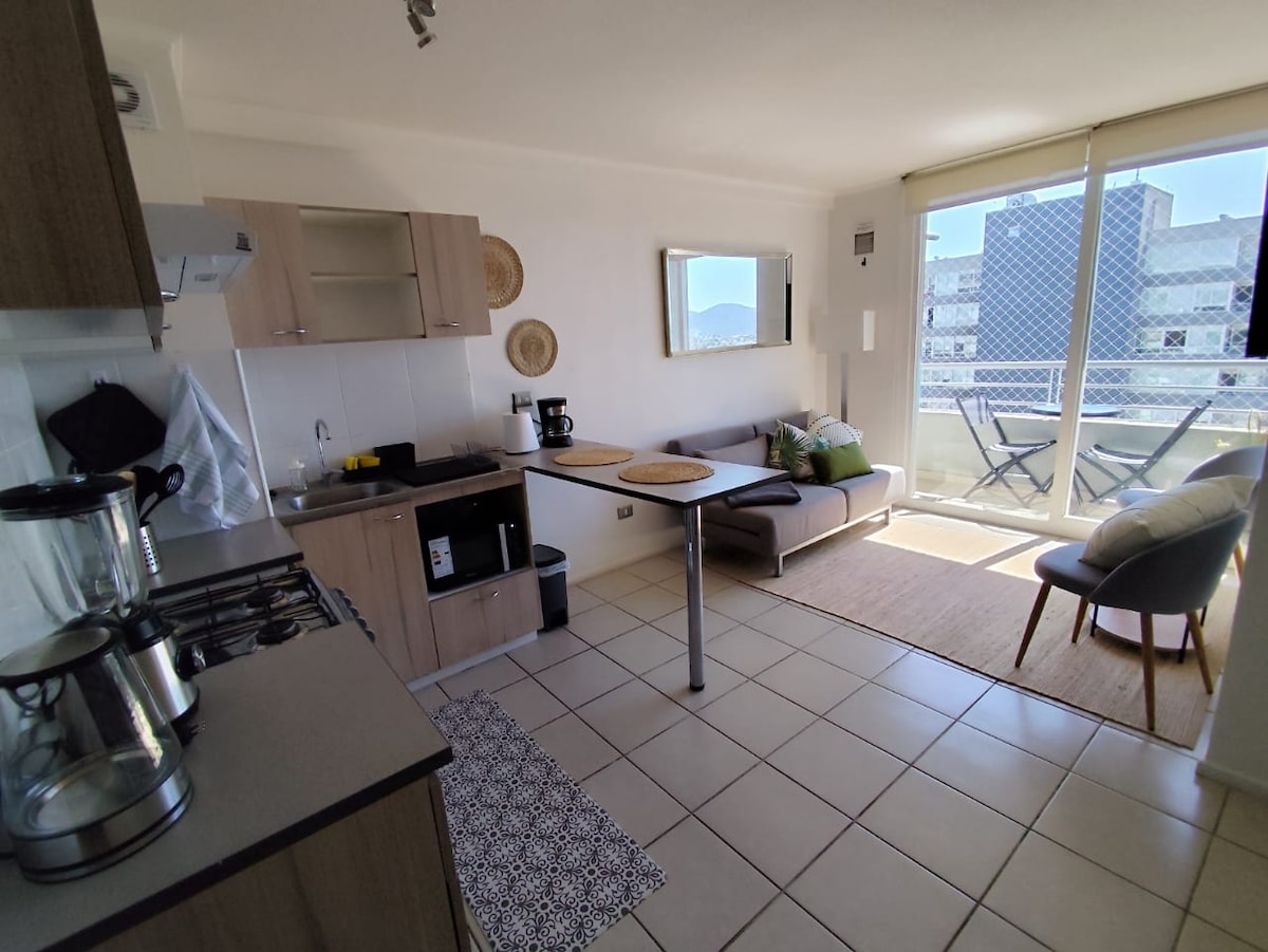 A modern kitchen area features sleek cabinetry and a compact dining table. A comfortable sofa is positioned near a window, allowing for natural light to fill the space. The flooring is tiled, enhancing the room's clean and welcoming vibe.