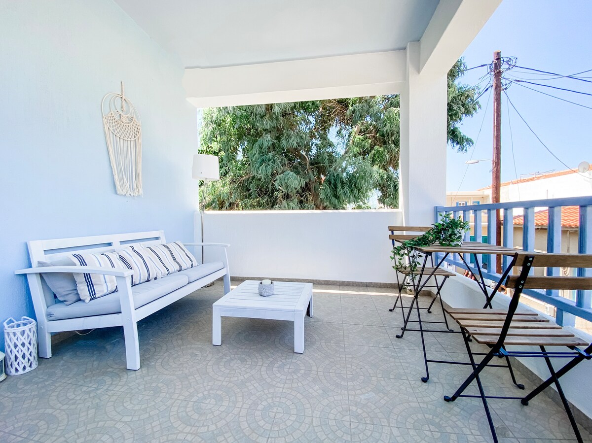 A spacious balcony features white outdoor furniture, including a couch and a coffee table. Two bistro-style chairs complement the setting, surrounded by greenery and natural light. The floor showcases a decorative tile pattern, enhancing the inviting atmosphere.