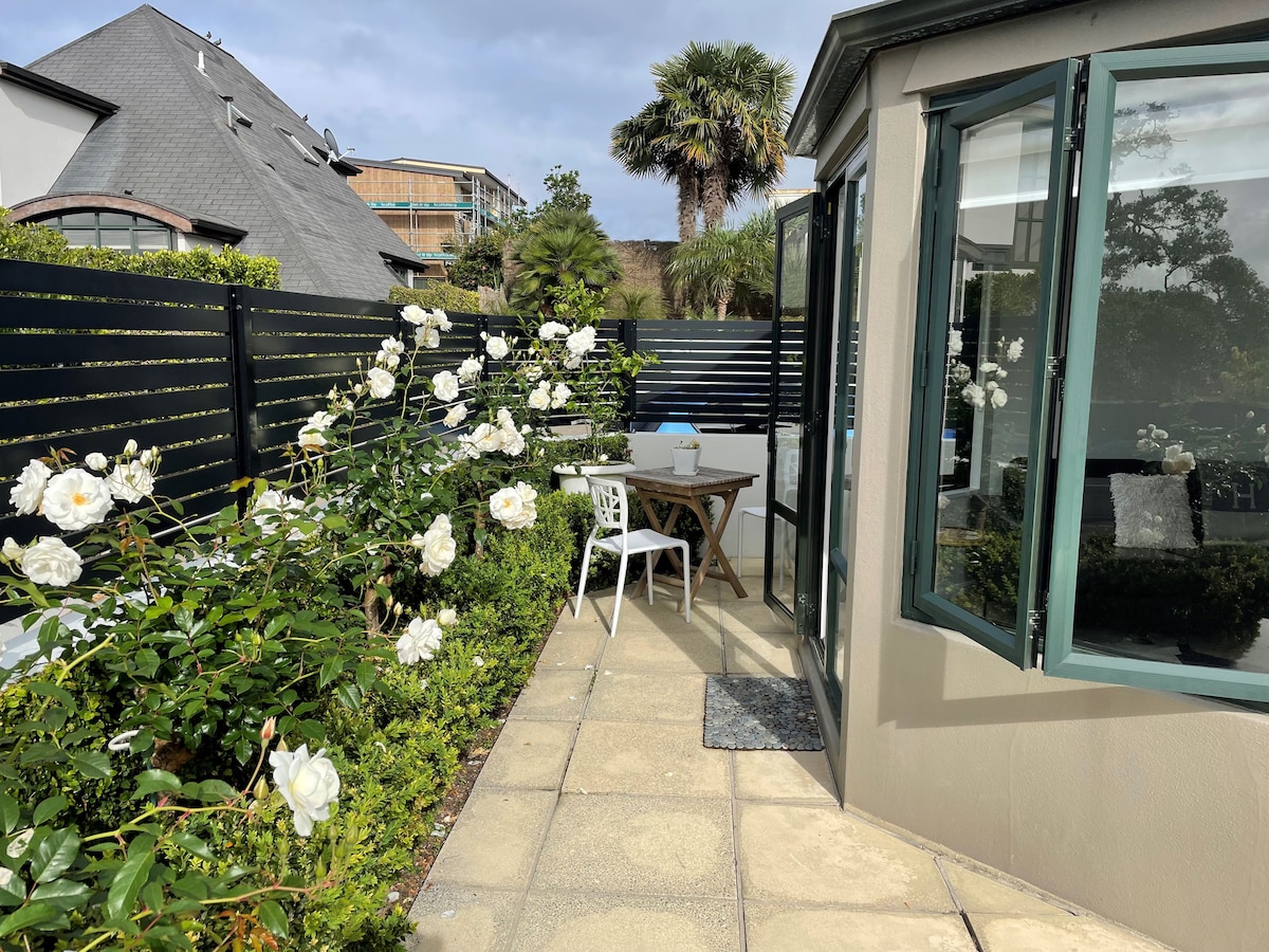 An outdoor area is depicted, featuring a small round table with two chairs under a canopy of white roses. Large windows provide access to the room, and a pathway of light-colored tiles leads through the garden. Lush greenery adds a welcoming touch.