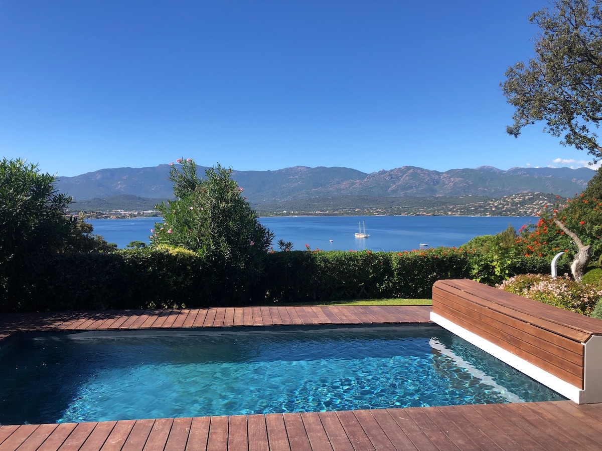 A private heated pool is surrounded by wooden decking and lush greenery, offering an unobstructed view of the sea and the distant mountains. Clear blue skies enhance the serene setting, with the peaceful waters reflecting the natural beauty of the landscape.