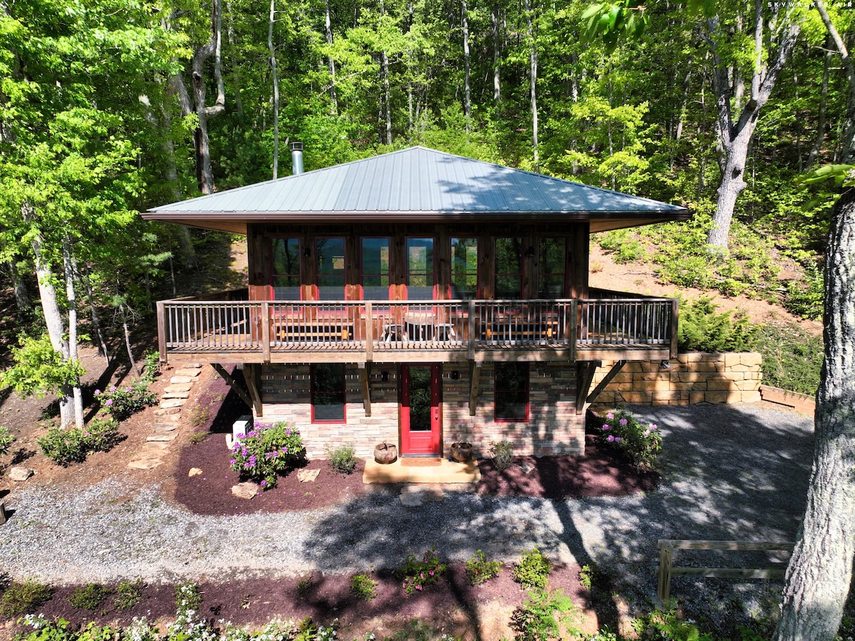 A two-story cabin is set among lush greenery, featuring a large deck with sturdy railings. The exterior is constructed from natural materials, complemented by vibrant landscaping and a gravel pathway. Large windows invite natural light, emphasizing the cabin's harmonious connection to the surrounding woods.
