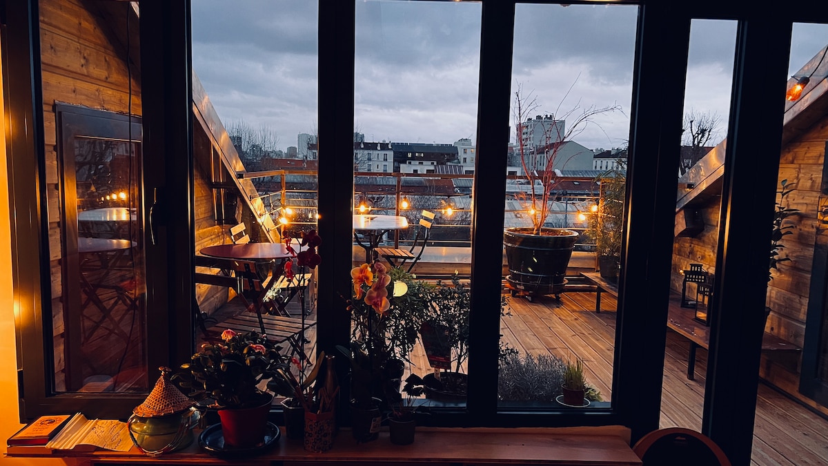 A rooftop terrace is showcased, surrounded by large glass windows. Various plants are visible on the deck, along with seating arrangements. Soft lighting enhances the evening ambiance, revealing a view of the urban skyline beyond.