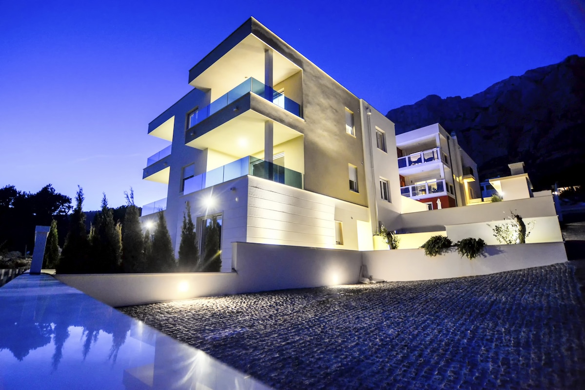 A modern multi-story building is illuminated against a twilight sky, highlighting its architectural design. The façade features a mix of light-colored walls and large balconies. Lush greenery and soft lighting enhance the surrounding area, creating a serene atmosphere.