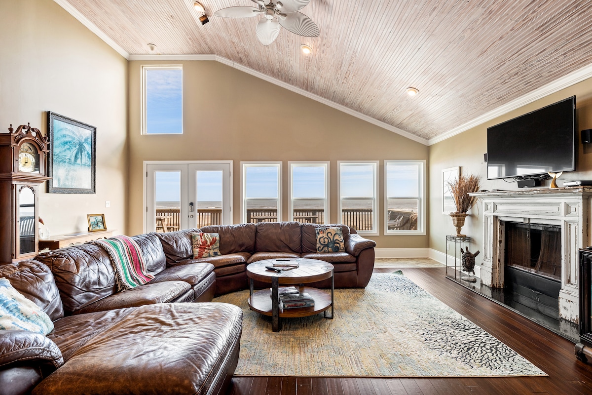 The spacious living area features a large sectional sofa with colorful throw pillows and a round coffee table at its center. Natural light fills the room through multiple windows, offering ocean views. A mounted television and decorative fireplace enhance the cozy, inviting ambiance.