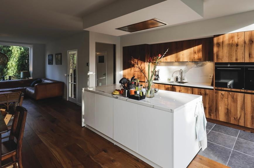 A modern kitchen is presented with a central island featuring a sleek white surface, complemented by wooden cabinetry. Stainless steel appliances are situated along the wall, and natural light filters through nearby windows, creating an inviting cooking space. A living area is visible in the background.