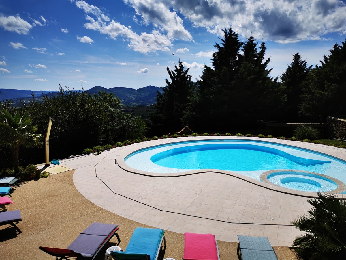 An inviting outdoor space features a uniquely shaped pool surrounded by a spacious patio. Lounge chairs in various colors are arranged around the pool, offering relaxation options. Lush greenery and distant mountains create a serene backdrop under a sky with scattered clouds.