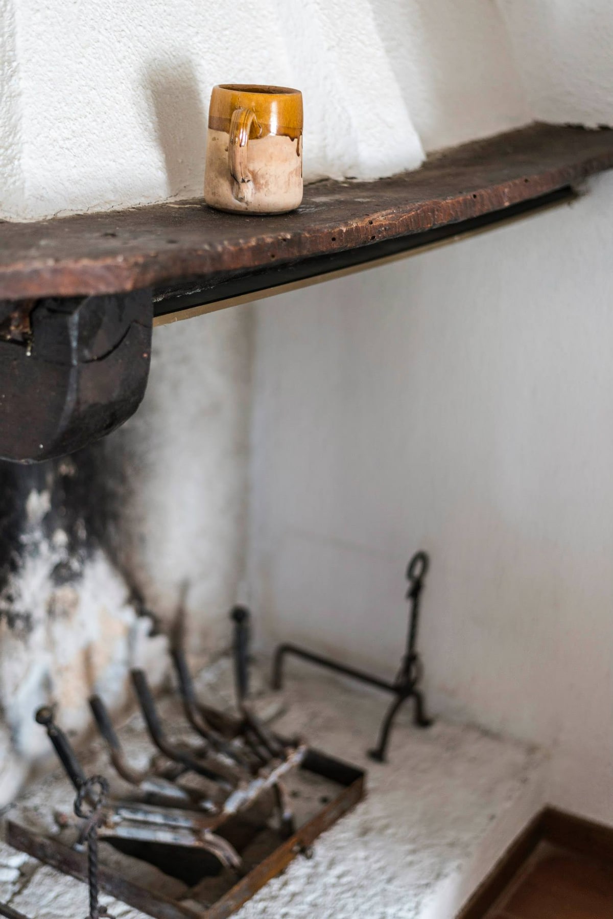 A traditional fireplace is situated in the corner of the room, featuring a rustic wooden mantle. A simple ceramic mug rests on the mantle above the whitewashed stone hearth, where iron tools are arranged neatly.