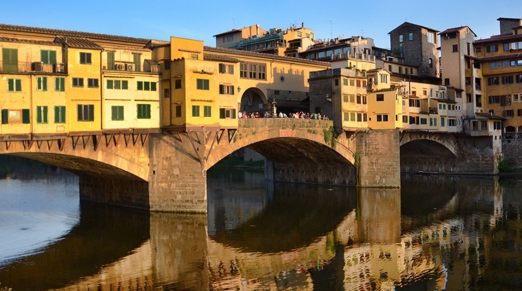 A historic bridge is visible, featuring a series of yellow and green buildings along its length. The structure arches gracefully over a calm river, with reflections mirrored in the water below. People can be seen enjoying the space on the bridge.