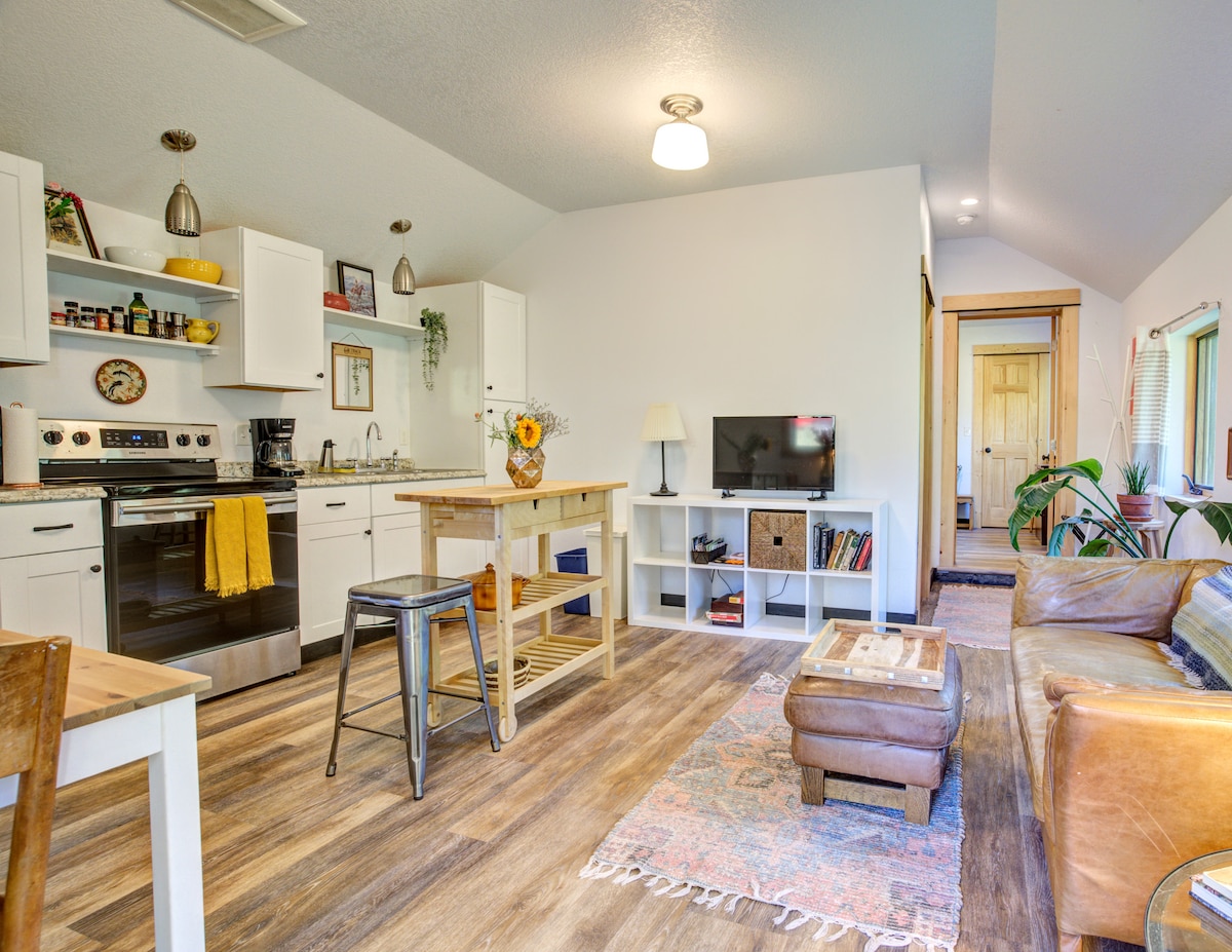 An open living space features modern kitchen cabinetry and a dining area with a wooden table. Comfortable seating is arranged near a television, with warm lighting illuminating the room. Decorative plants and a colorful area rug add a cozy touch to the inviting atmosphere.