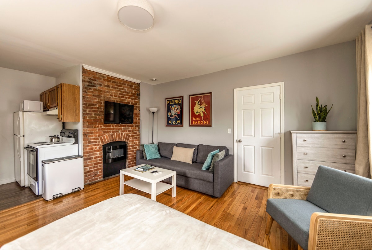 The spacious studio apartment features a cozy seating area with a gray sofa and a coffee table. A restored brick fireplace stands prominently, complemented by a wall-mounted TV. The kitchen area is visible, equipped with appliances, and light streams in through a large window.