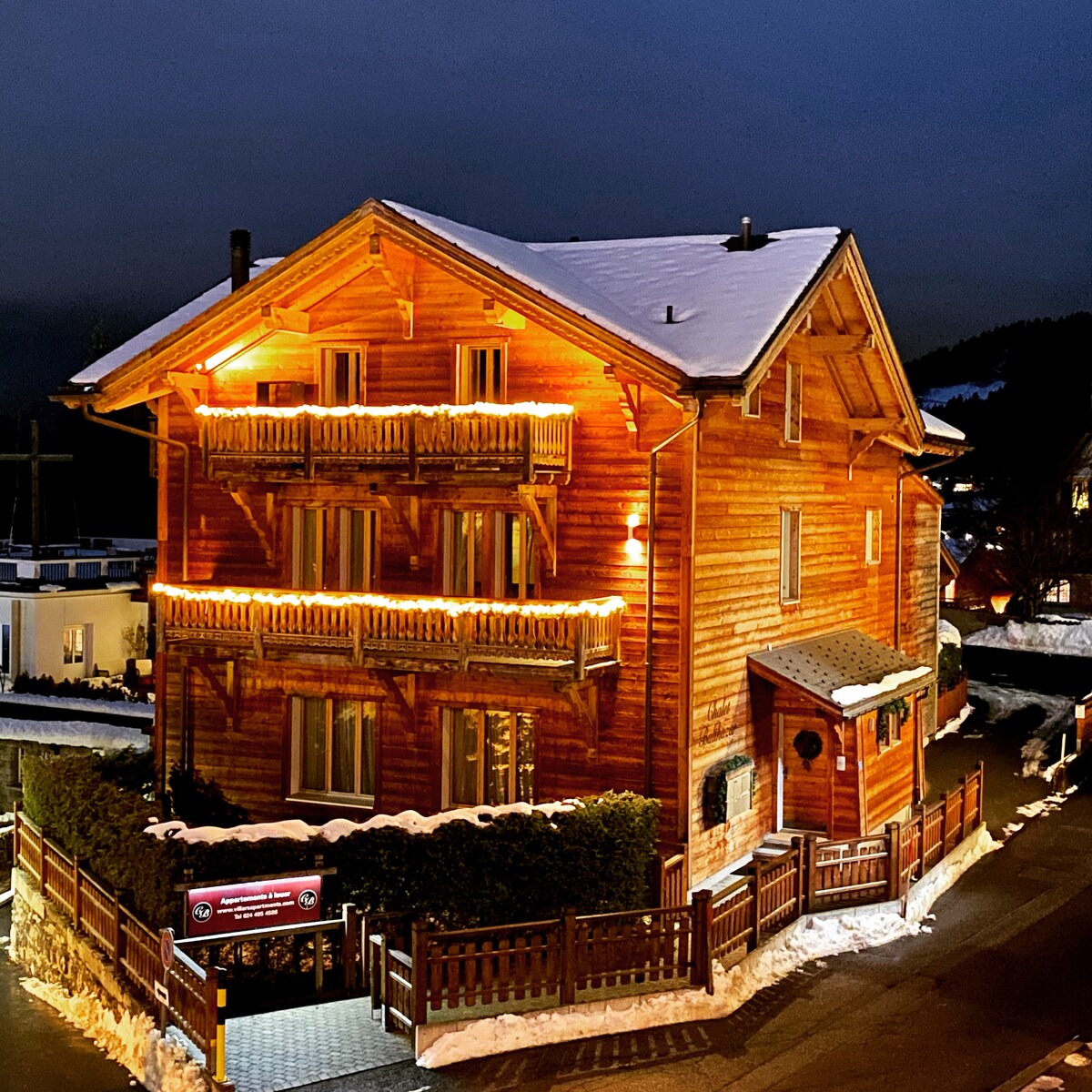 The chalet features a stunning wooden exterior adorned with warm lighting, creating a welcoming ambiance against the evening sky. Balconies extend from the upper levels, surrounded by snow-covered landscaping and a sturdy wooden fence that defines the property.