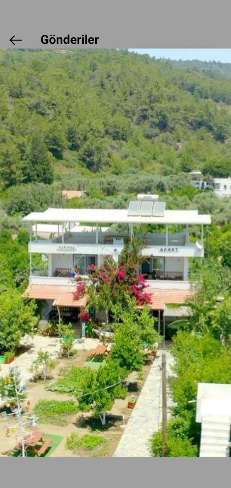 A multi-story building is surrounded by lush greenery, featuring blooming flowers and trees in the foreground. A terrace is visible on the upper level, and outdoor seating areas are situated in the garden below, offering a tranquil environment.