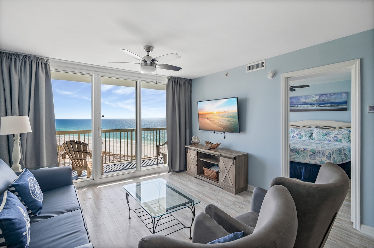 The living area features a spacious seating arrangement with a blue sofa and comfortable chairs, paired with a glass coffee table. A flat-screen TV is mounted on the wall. Large sliding glass doors lead to a balcony, offering unobstructed views of the beach and Gulf of Mexico.