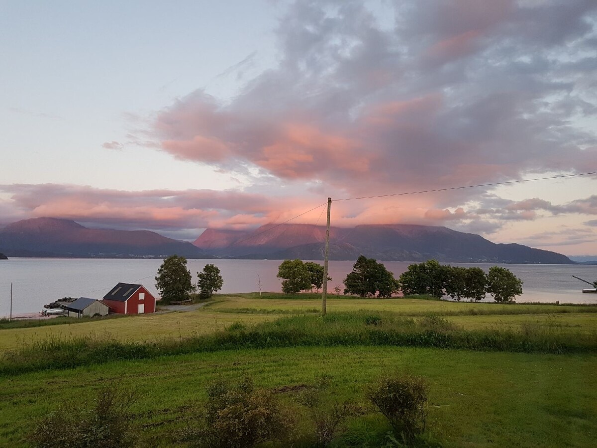 A scenic view of the Fjord is presented, with mountains in the background under a pastel sky. Green fields stretch towards the waterfront, while a red barn and a few trees are visible on the left, enhancing the natural beauty of the surroundings.