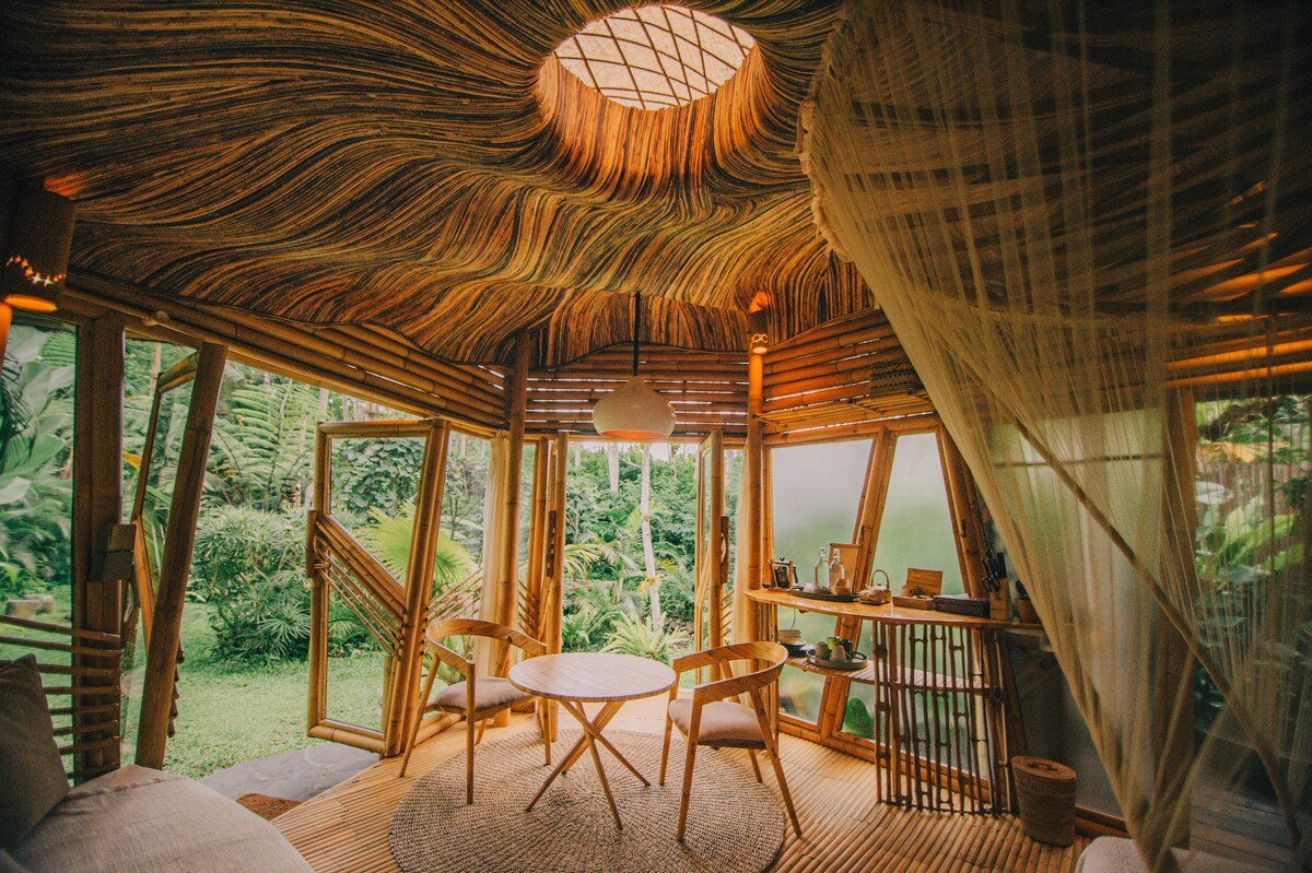 The bamboo interior features a woven ceiling that resembles natural waves, allowing light to filter in from a circular skylight. A round table with two chairs is set in a cozy corner, with shelves displaying thoughtfully arranged items, and large windows providing views of the surrounding greenery.