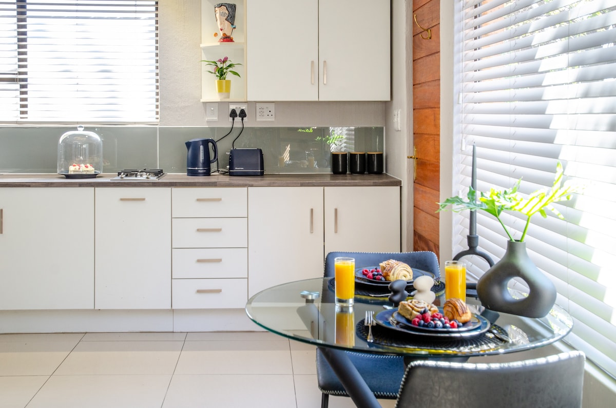 An open-plan kitchen and dining area feature a glass dining table set for two, complete with plates of fruit, pastries, and glasses of juice. A kettle and modern appliances are visible on the countertop, while natural light streams through the partially open window.