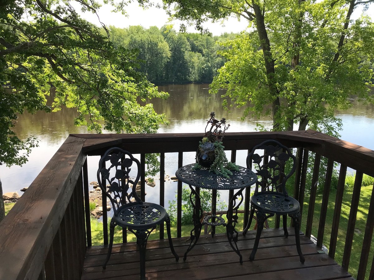 A wooden deck features a small round table surrounded by two ornate metal chairs. Green foliage frames the view of the Flambeau River, with calm waters reflecting the surrounding trees under a bright sky.