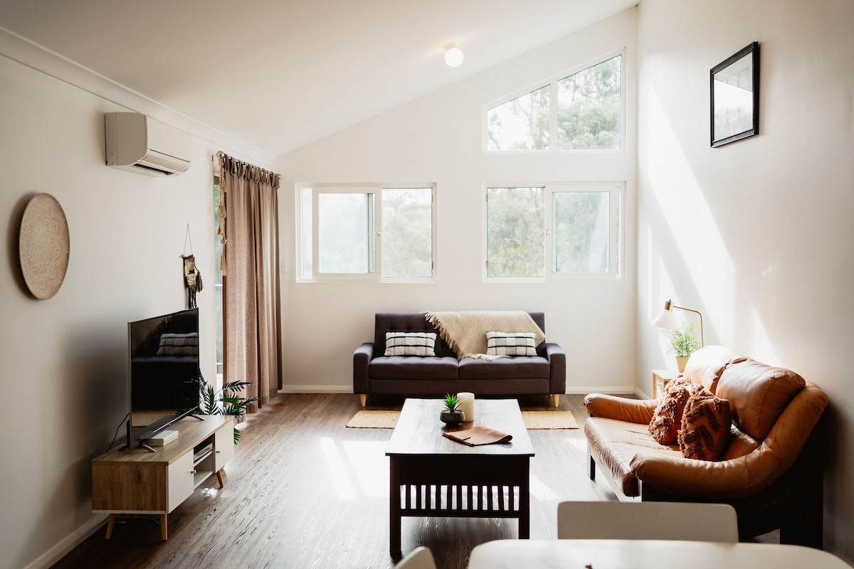 A bright living area is illuminated by natural light streaming through large windows. A brown leather sofa is paired with a dark wooden coffee table, while a modern TV unit holds a television. A cozy throw blanket is draped over a gray fabric sofa, enhancing the relaxed ambiance.