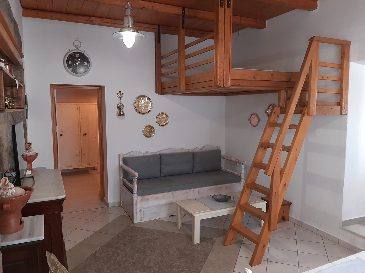 An interior space features a wooden staircase leading to a raised sleeping area. A light-colored sofa is placed against the wall, accompanied by a small coffee table. Decorative plates are displayed on the wall, while tiled flooring adds a classic touch to the setting.