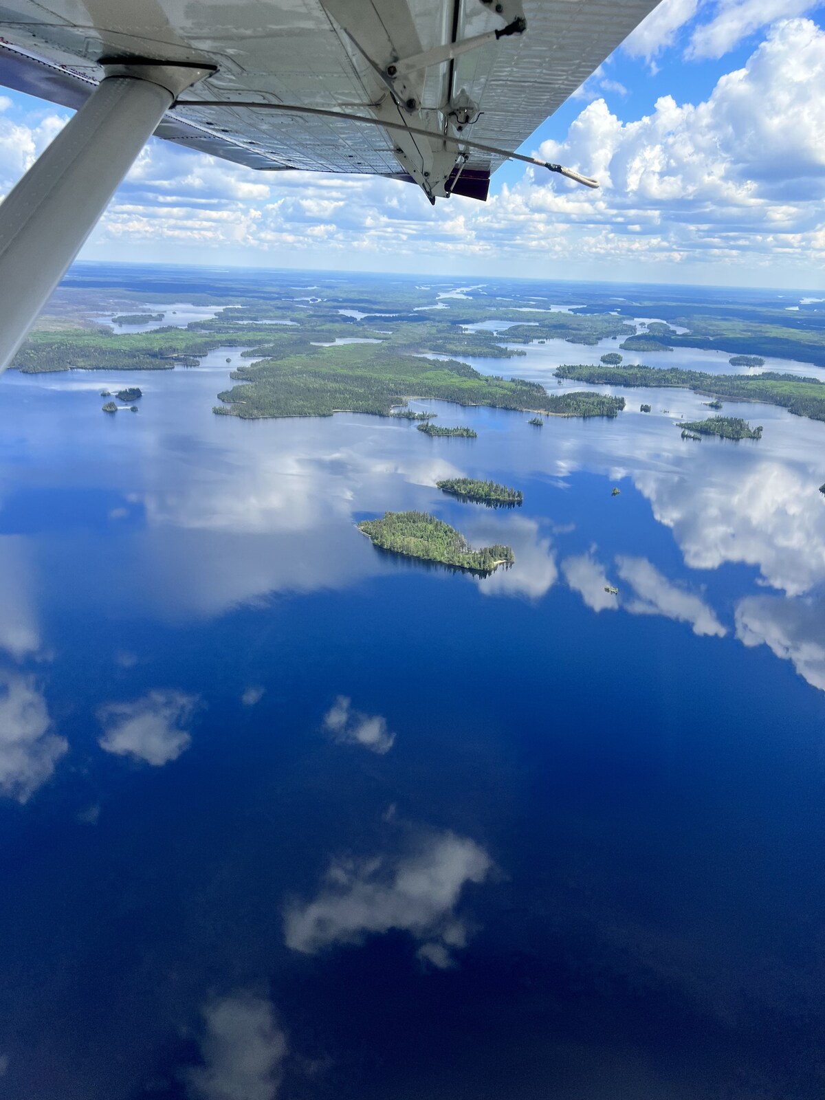 An aerial view captures calm, reflective waters of Melgurd Lake, dotted with lush green islands and bordered by dense forests. The sky is expansive, featuring fluffy white clouds casting soft shadows on the surface below. The serenity of the landscape highlights the cabin's isolated location.