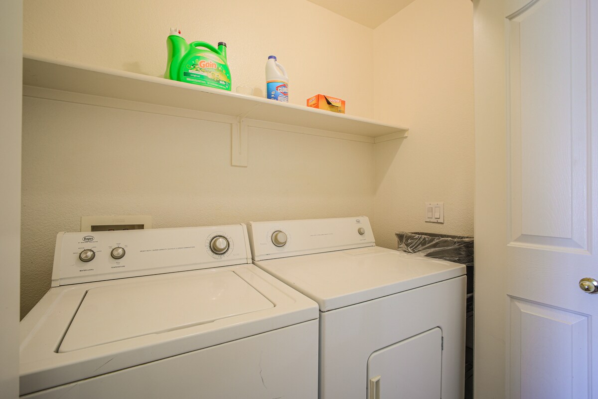 A dedicated laundry area is equipped with a washing machine and dryer side by side. Above, shelves hold cleaning supplies and laundry essentials, while a small waste bin is located to the side. The walls are painted in a soft, neutral tone.