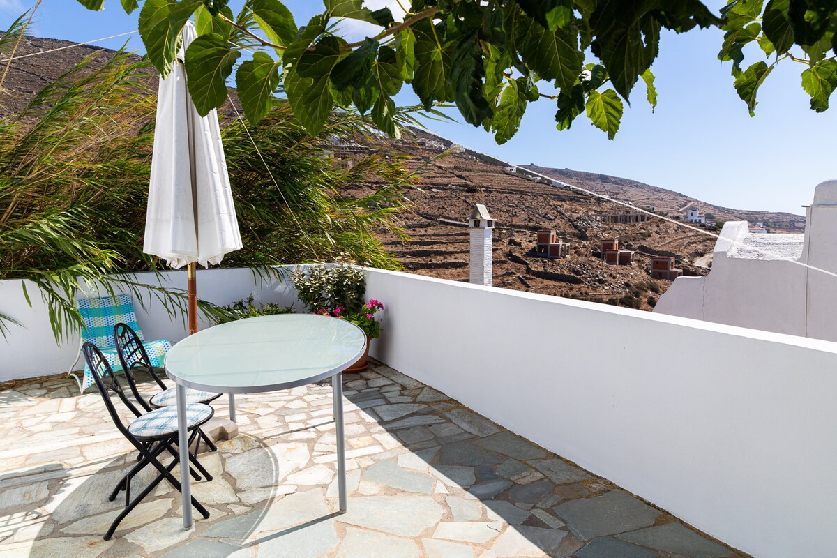 An outdoor terrace features a round glass table surrounded by four chairs, shaded by a vibrant umbrella. A view of the natural landscape and distant hills is visible, complemented by greenery along the perimeter.