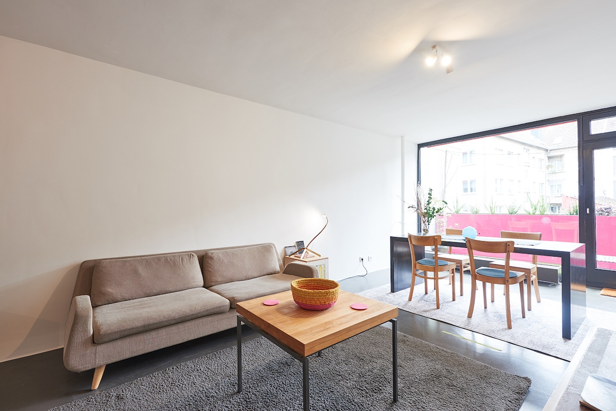 A modern living area is showcased, featuring a light-colored sofa and a wooden coffee table with a decorative bowl. A dining area with wooden chairs is visible, alongside an expansive window that allows natural light to illuminate the space. A soft rug covers the floor.