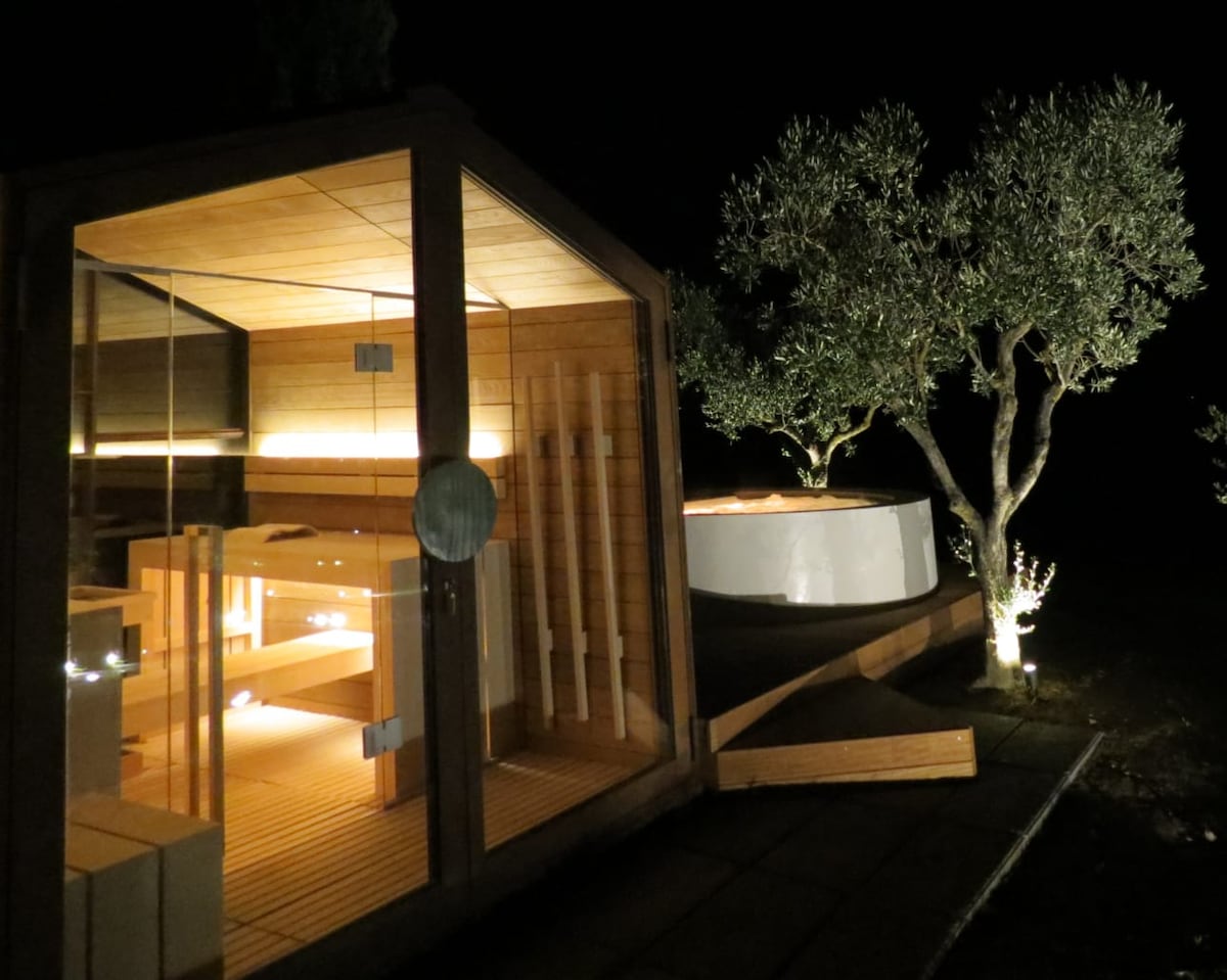 A modern sauna built with warm wooden panels is illuminated softly at night, accompanied by a circular hot tub. Olive trees surrounding the area are subtly lit, creating a serene outdoor relaxation space.
