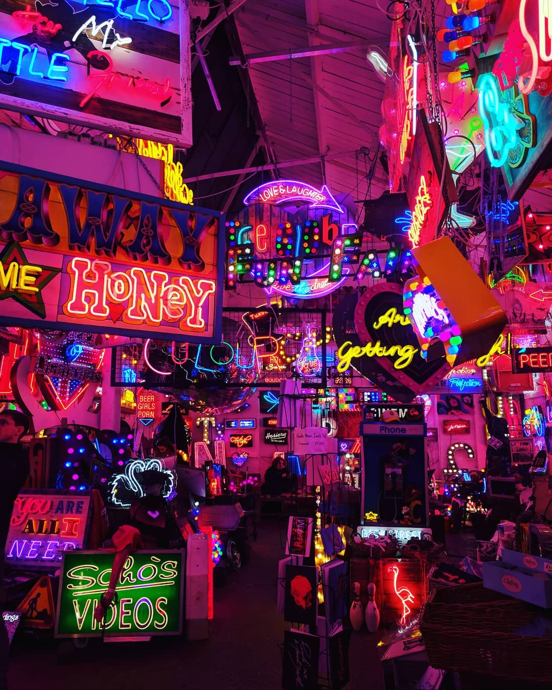 A vibrant display of neon signs fills the space with a variety of colors and designs. The eclectic assortment illuminates both the ceiling and walls, creating an energetic atmosphere. Various phrases and images are depicted, contributing to a lively visual experience.