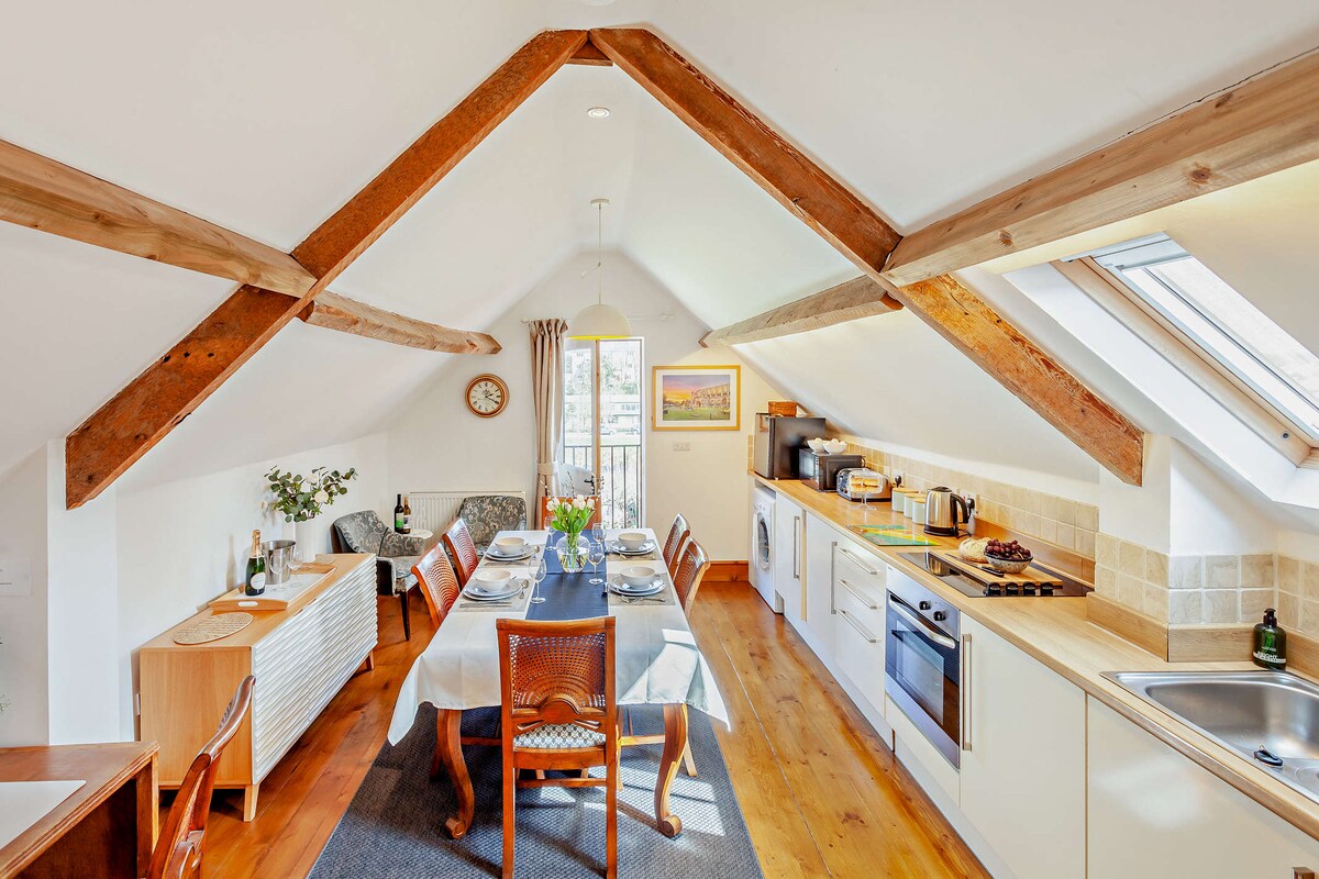 The kitchen and dining area feature exposed wooden beams and large windows allowing natural light. A wooden dining table with seating for six is set with tableware. Nearby, modern appliances, including a stove and microwave, are neatly arranged along the countertops.