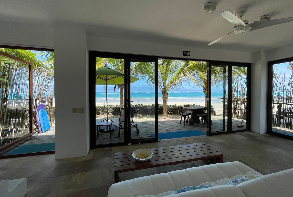 The spacious living area showcases large glass doors that frame scenic beach views, allowing natural light to fill the room. A white sectional sofa is positioned near a wooden coffee table. Outside, a terrace hosts sun loungers and an umbrella amid swaying palm trees.