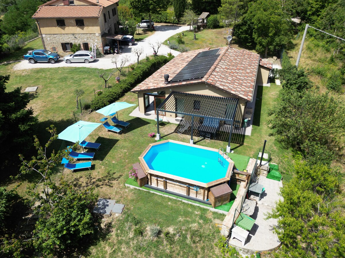 An aerial view of the property reveals a private garden with a vibrant blue pool surrounded by lounge chairs and umbrellas. A stone building can be seen in the background, nestled among lush greenery. Solar panels are visibly installed on the roof.