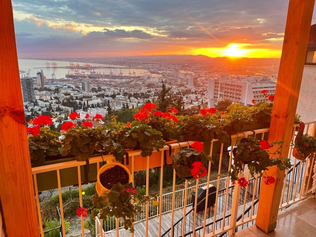 A balcony adorned with vibrant red flowers offers a stunning view of the sunset over the city, with the sea and harbor in the distance. The warm hues of the sky are reflected in the scene, creating a serene and picturesque atmosphere.