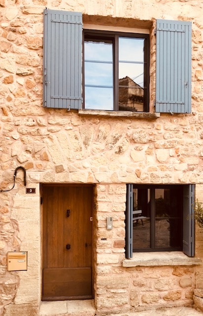The stone facade of a charming house is visible, featuring blue shutters and a wooden front door. A large window allows natural light to enter, reflecting the surroundings. The entrance is framed by two open windows, enhancing the welcoming appearance of the property.